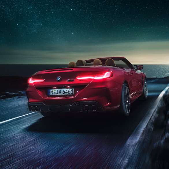 BMW M8 Competition Cabrio, tylny półprofil. BMW M8 Competition Cabrio w metalizowanym kolorze czerwieni Motegi z otwartym dachem, tylny półprofil, jadące w nocy drogą wzdłuż wybrzeża.