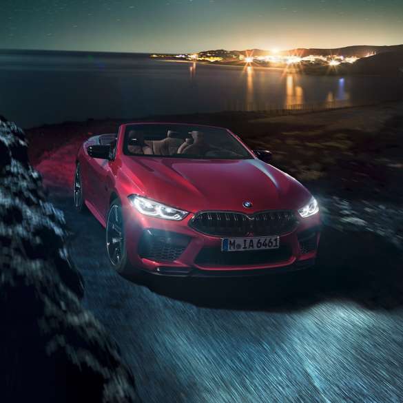 BMW M8 Competition Cabrio, przedni półprofil. BMW M8 Competition Cabrio w metalizowanym kolorze czerwieni Motegi z otwartym dachem, przedni półprofil, jadące w nocy drogą wzdłuż wybrzeża.