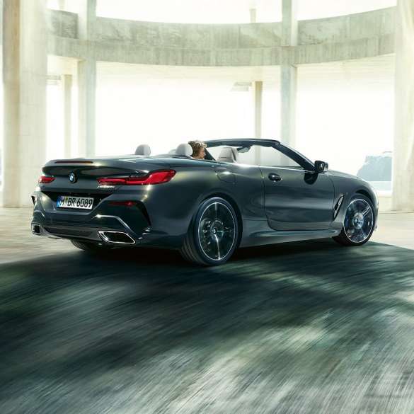 BMW M850i xDrive Cabrio, tylny półprofil. BMW M850i xDrive Cabrio w metalizowanym kolorze szarego drawitu z otwartym dachem, tylny półprofil, jadące przez betonową budowlę.