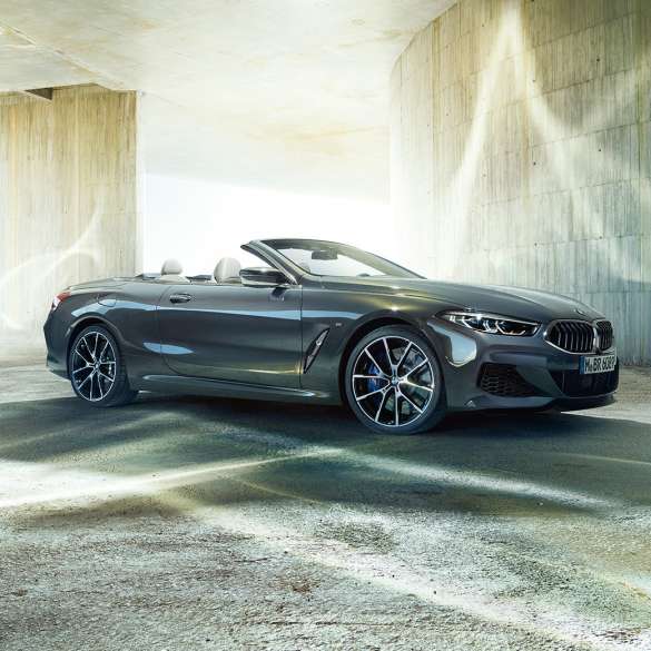 BMW M850i xDrive Cabrio, półprofil z boku. BMW M850i xDrive Cabrio w metalizowanym kolorze szarego drawitu z otwartym dachem, półprofil z boku, stojące przed betonową budowlą.