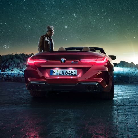 BMW M8 Competition Cabrio, widok z tyłu. BMW M8 Competition Cabrio w metalizowanym kolorze czerwieni Motegi z otwartym dachem, widok z tyłu, stojące wraz z mężczyzną pod rozgwieżdżonym niebem.