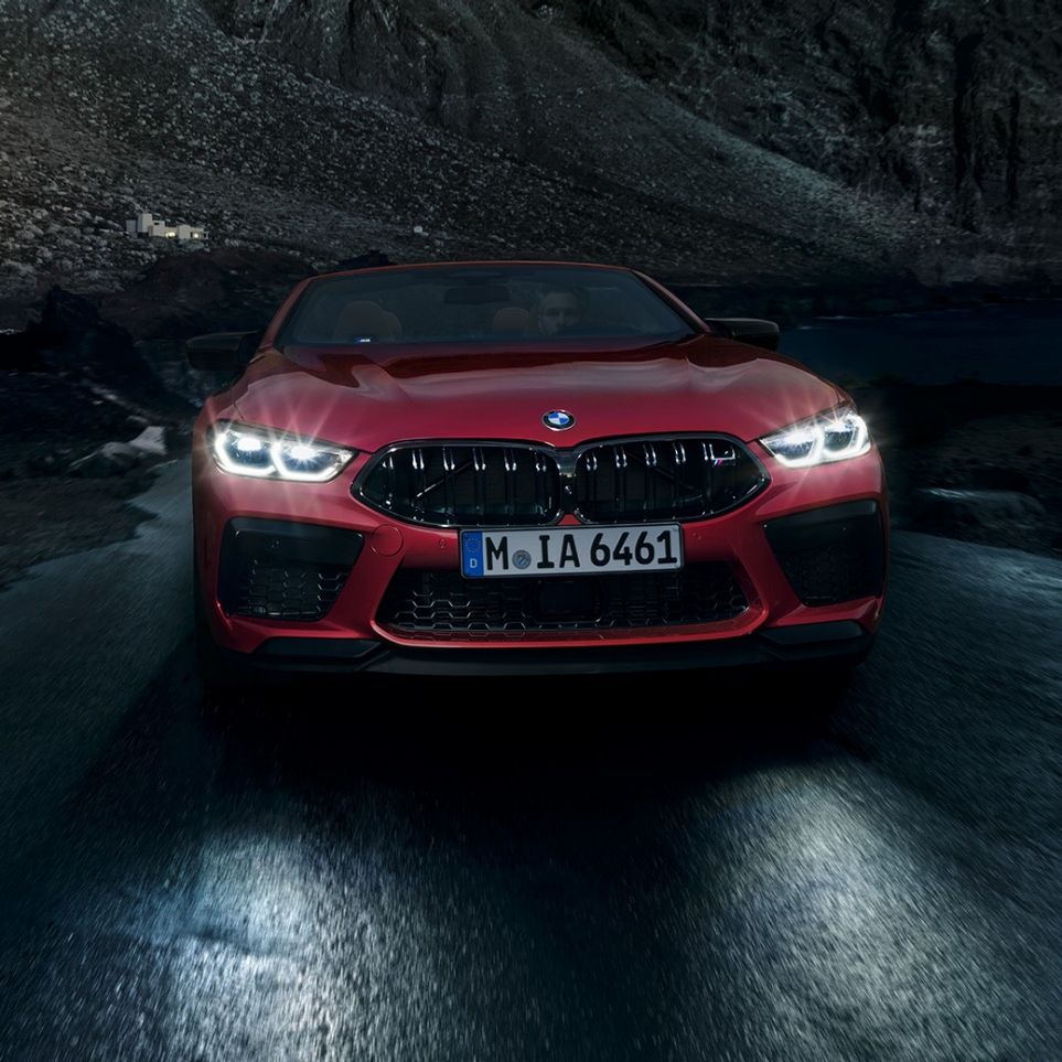 BMW M8 Competition Cabrio, widok z przodu. BMW M8 Competition Cabrio w metalizowanym kolorze czerwieni Motegi ze świecącymi reflektorami, widok z przodu, jadące w nocy.