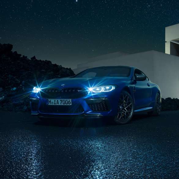 BMW M8 Competition Coupé, przedni półprofil. BMW M8 Competition Coupé w metalizowanym kolorze niebieskim Marina Bay ze świecącymi reflektorami LED, przedni półprofil w bezruchu, w nocy na tle willi.