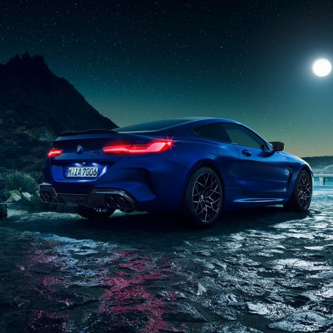 BMW M8 Competition Coupé, tylny półprofil. BMW M8 Competition Coupé w metalizowanym kolorze niebieskim Marina Bay ze świecącymi reflektorami LED, tylny półprofil w bezruchu, przy księżycu w pełni.