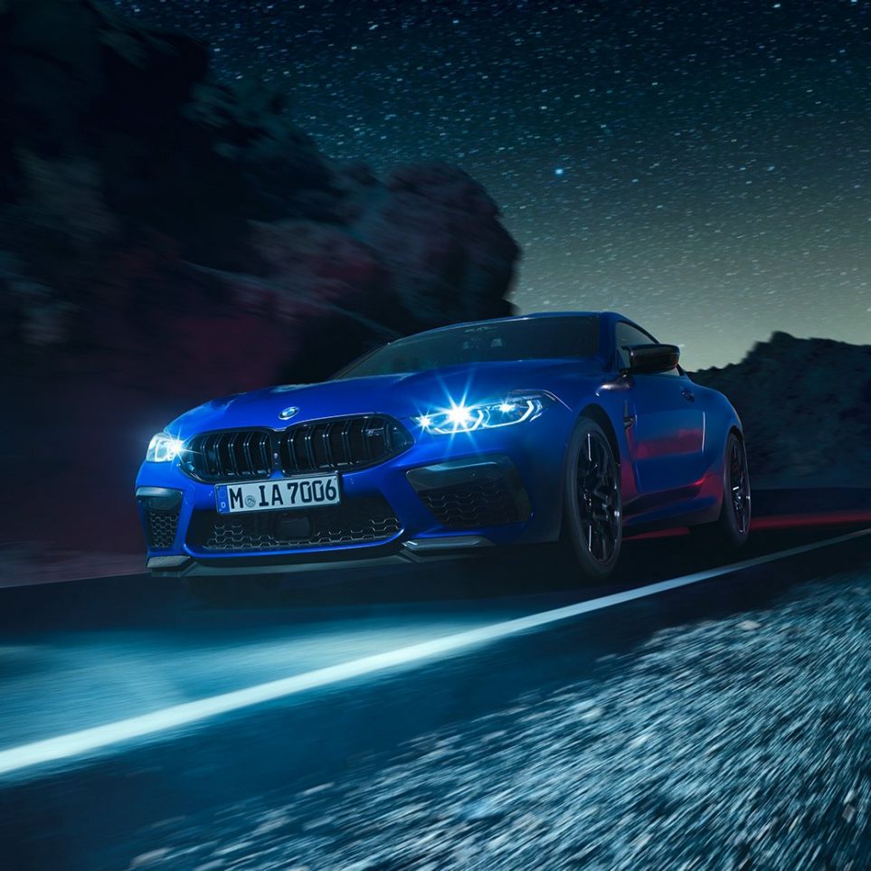 Jadące BMW M8 Competition Coupé, przedni półprofil. BMW M8 Competition Coupé w metalizowanym kolorze niebieskim Marina Bay ze świecącymi reflektorami LED, przedni półprofil, jadące w nocy.