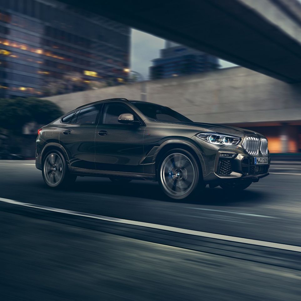 BMW X6 podczas jazdy. BMW X6, ujęcie w półprofilu od przodu auta jadącego na tle scenerii miejskiej.
