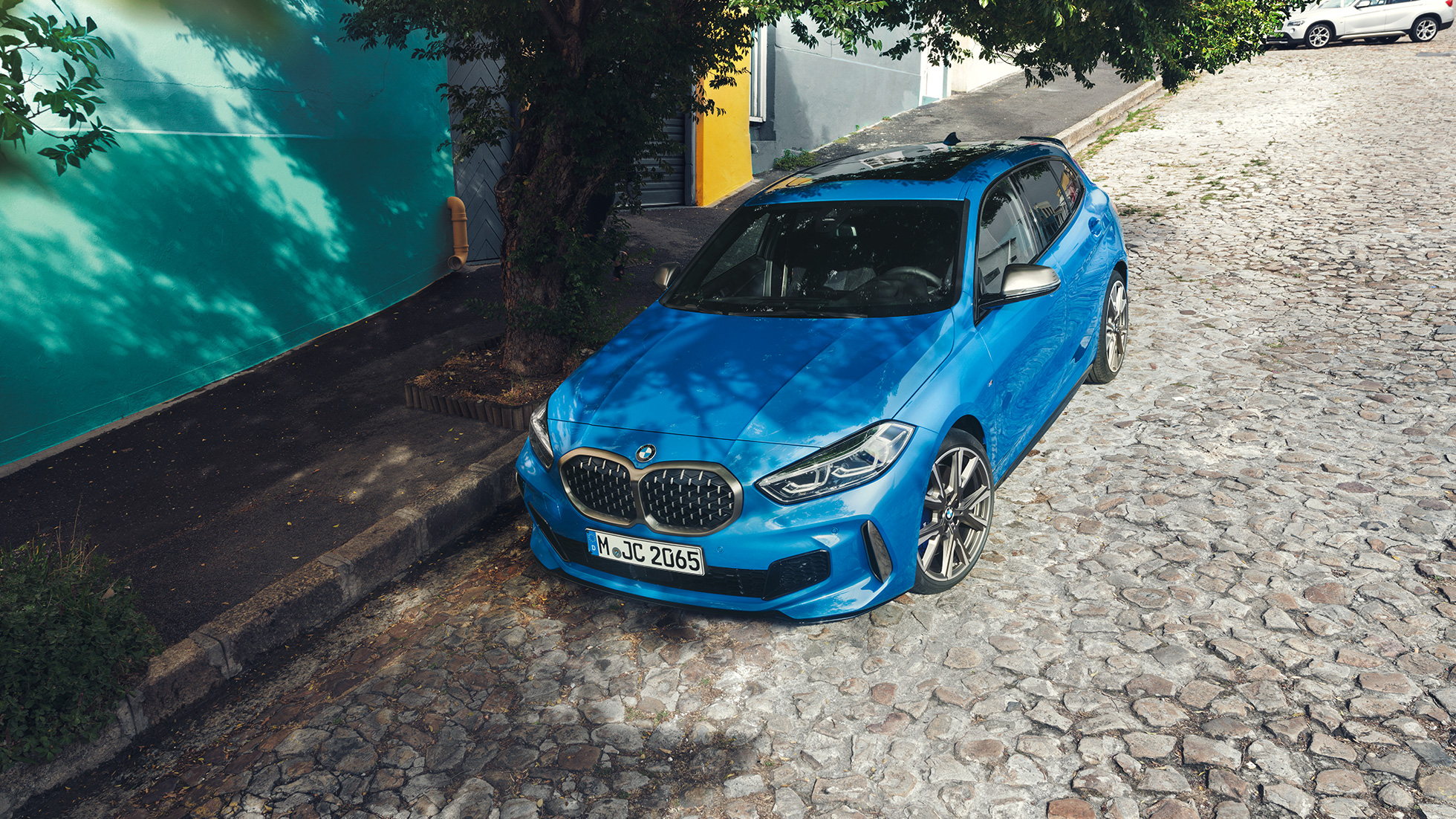 BMW serii 1 na poboczu drogi. BMW serii 1 w metalizowanym kolorze niebieskim Misano, przedni półprofil, zaparkowane na poboczu drogi z lotu ptaka.