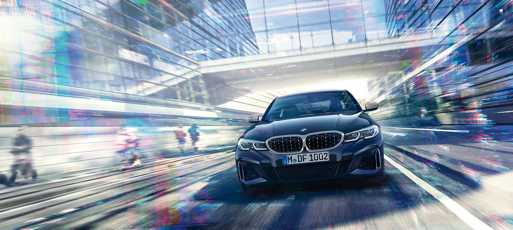 BMW serii 3 Limuzyna, widok z przodu BMW M340i xDrive. Widok z przodu na jadące BMW M340i xDrive Limuzynę pomiędzy szklanymi budynkami.