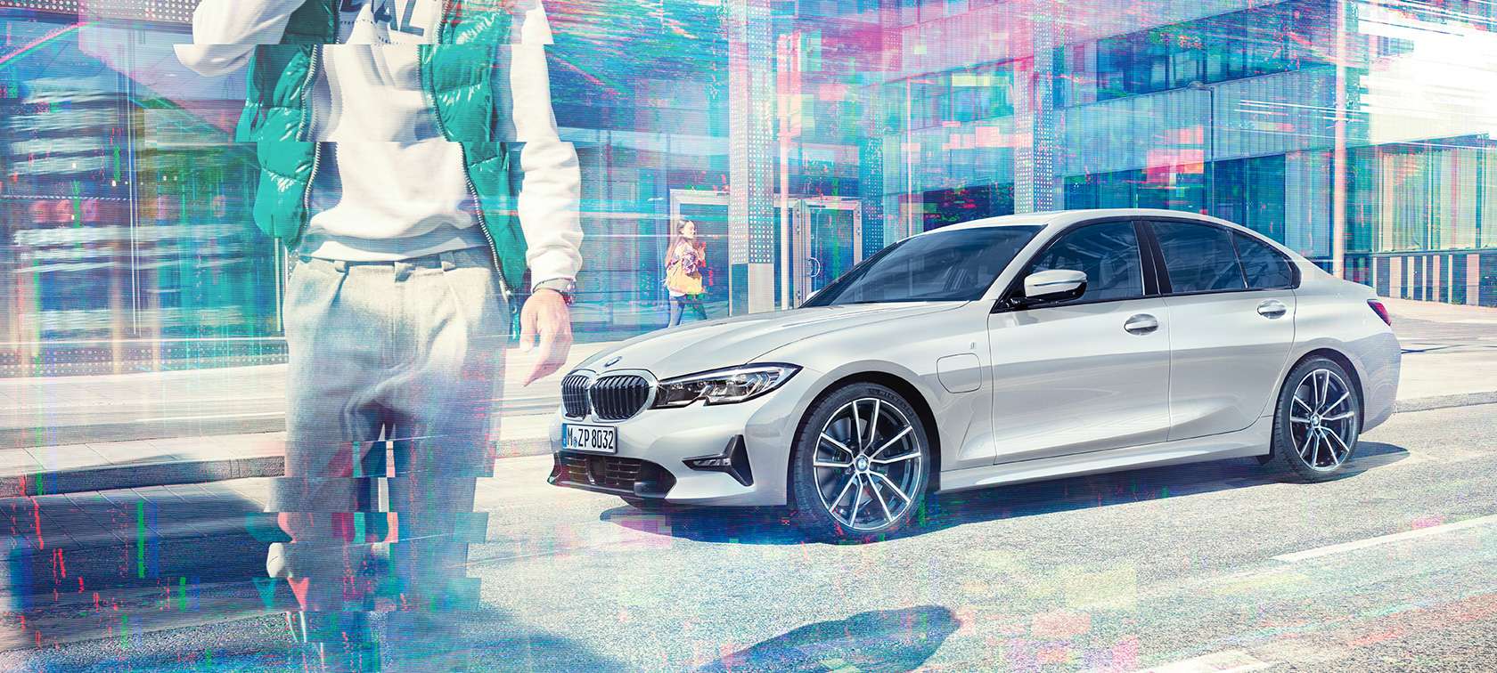 BMW serii 3 Limuzyna, widok z boku BMW 330e iPerformance. Ujęcie trzy-czwarte na bok stojącego BMW 330e Limuzyna iPerformance przed szklanym budynkiem z przechodzącymi pieszymi.