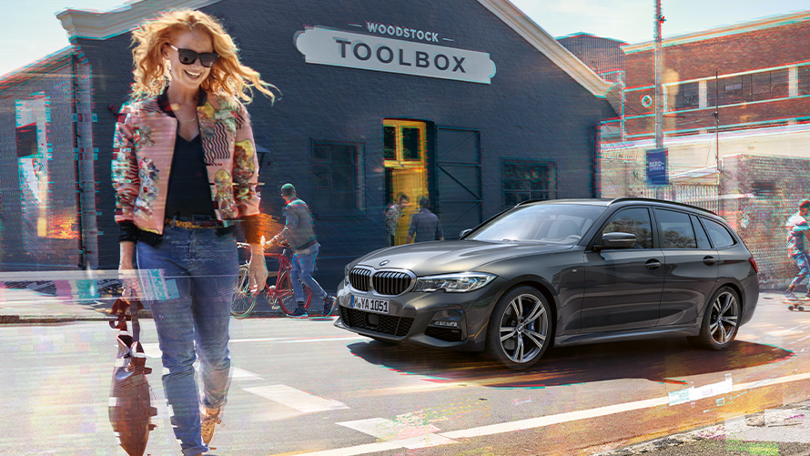 BMW serii 3 Touring (G21), przedni półprofil na ulicy z kobietą przed sklepem stolarskim Kobieta przechodzi przez jezdnię przed BMW serii 3 Touring (G21) i sklepem stolarskim.