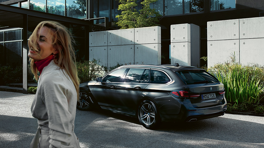 Kobieta BMW serii 5 Touring G31 LCI szary Sophisto z brylantowym efektem metalizowany widok z boku 7/8 Kobieta przed zaparkowanym BMW serii 5 Touring G31 modernizacja 2020 szary Sophisto z brylantowym efektem metalizowany widok z boku 7/8