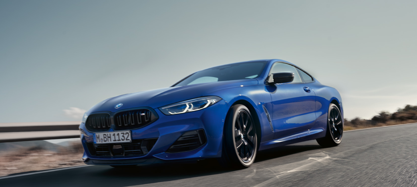 BMW serii 8 Coupé G15 LCI modernizacja 2022 BMW M850i xDrive niebieski Portimao M metalizowany widok z przodu ujęcie z dołu podczas jazdy BMW serii 8 Coupé G15 LCI modernizacja 2022 BMW M850i xDrive niebieski Portimao M metalizowany widok z przodu ujęcie z dołu podczas jazdy poza miastem na tle pagórkowatego krajobrazu