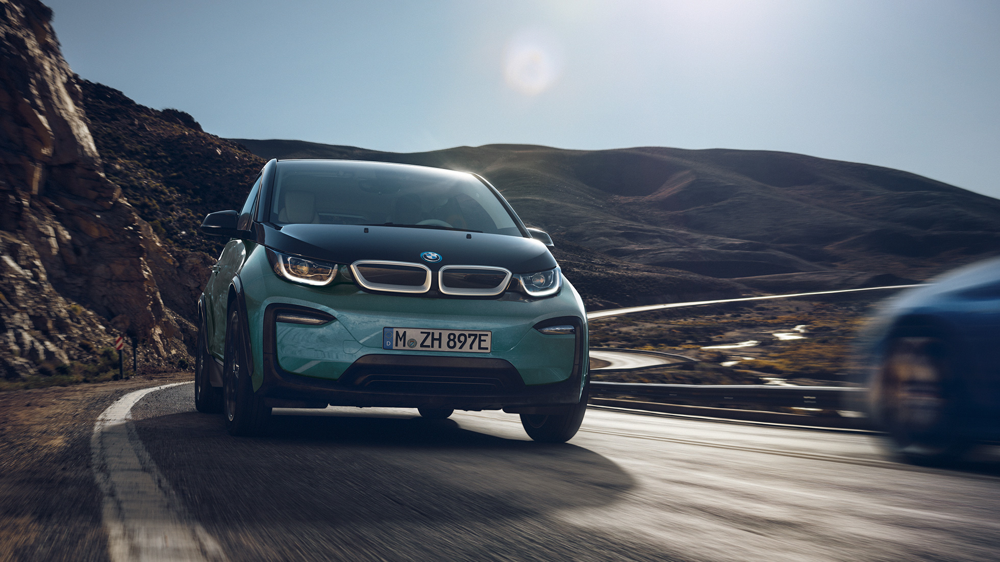 BMW i3 2018, widok z boku jadącego samochodu BMW i3 I01 2018, beż Jucaro z akcentami w metalizowanym kolorze mroźnym szarym, widok z boku jadącego samochodu na tle miasta