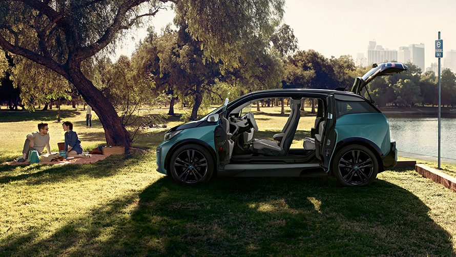 BMW i3 2018 w parku BMW i3 I01 2018, beż Jucaro z akcentami w metalizowanym kolorze mroźnym szarym, widok z boku z otwartymi drzwiami w parku
