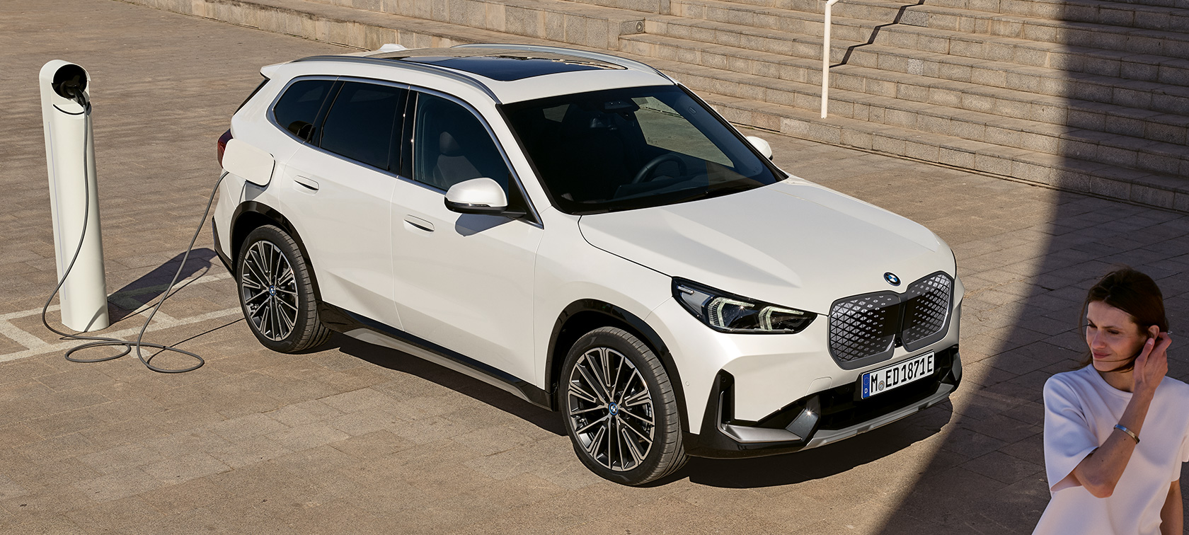 BMW iX1 U11 BEV widok z boku podczas ładowania BMW iX1 U11 BEV 2022 biel mineralna metalizowany boczny półprofil podczas ładowania schody