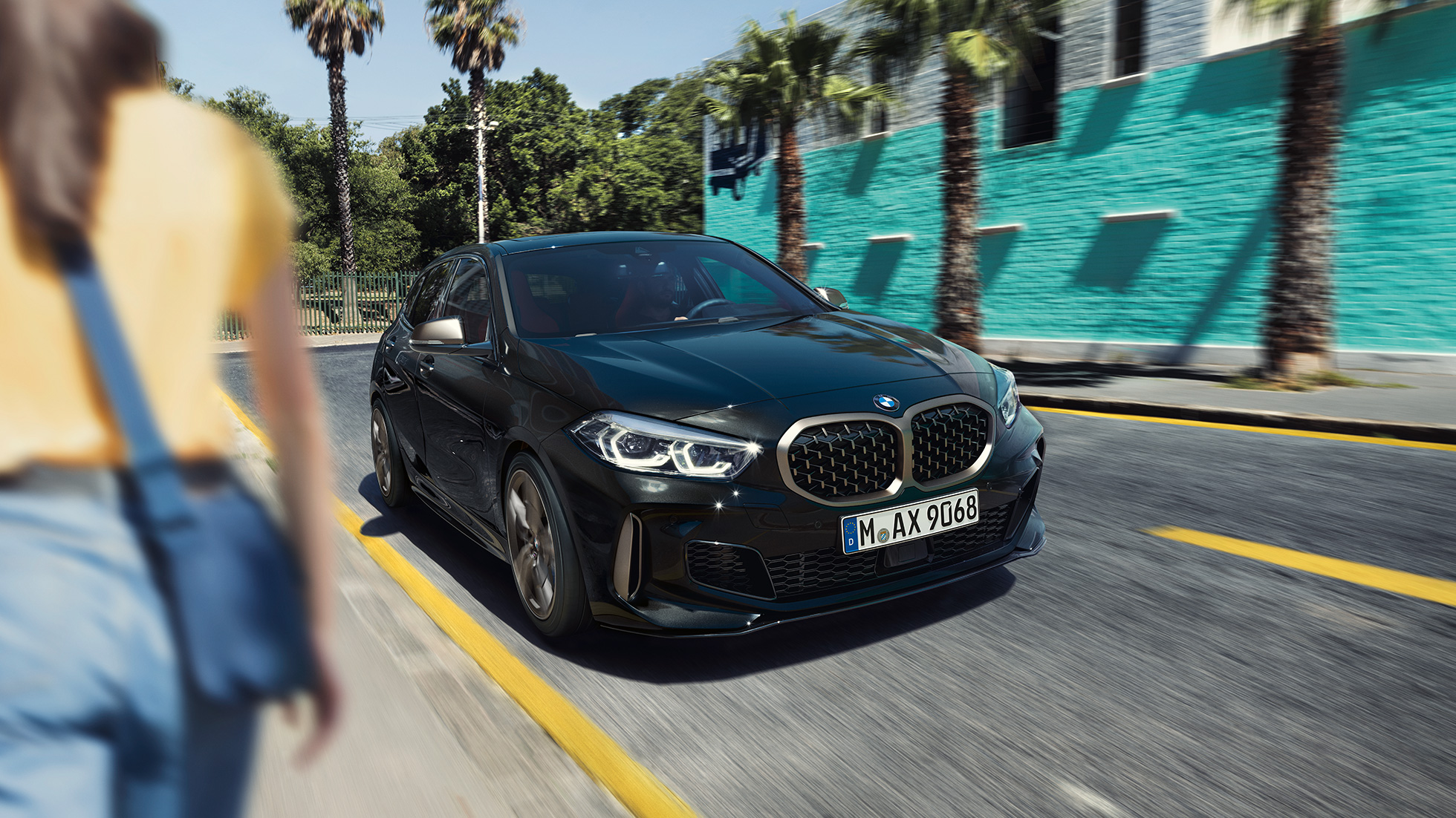 BMW M135i xDrive, przedni półprofil. BMW M135i xDrive w metalizowanym kolorze szafirowoczarnym, przedni półprofil podczas jazdy, z kobietą na pierwszym planie.