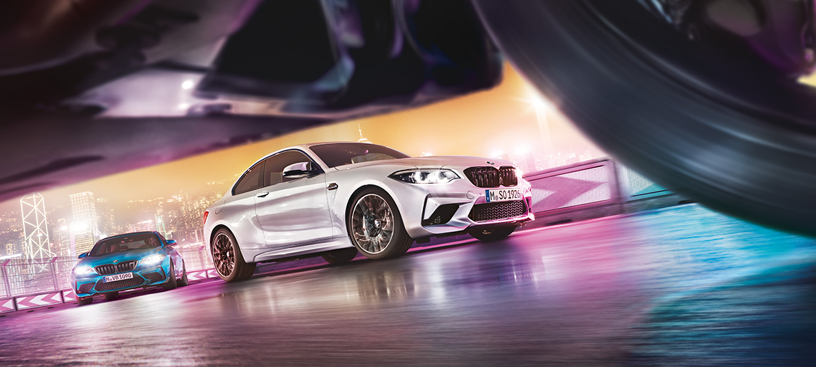 BMW M2 Competition na rozświetlonym asfalcie. Dwa BMW M2 Competition na torze wyścigowym w nocnym miejskim pejzażu z widokiem z perspektywy ulicy.