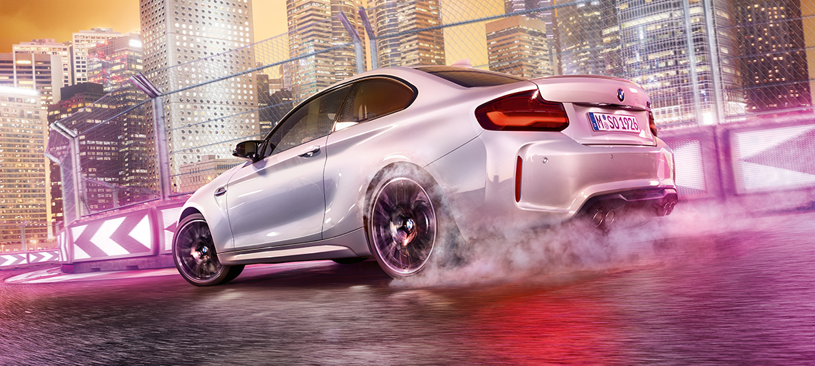 BMW M2 Competiotion w ruchu przyspieszenia. Białe BMW M2 Competition driftująca na torze wyścigowym przed nocną panoramą.