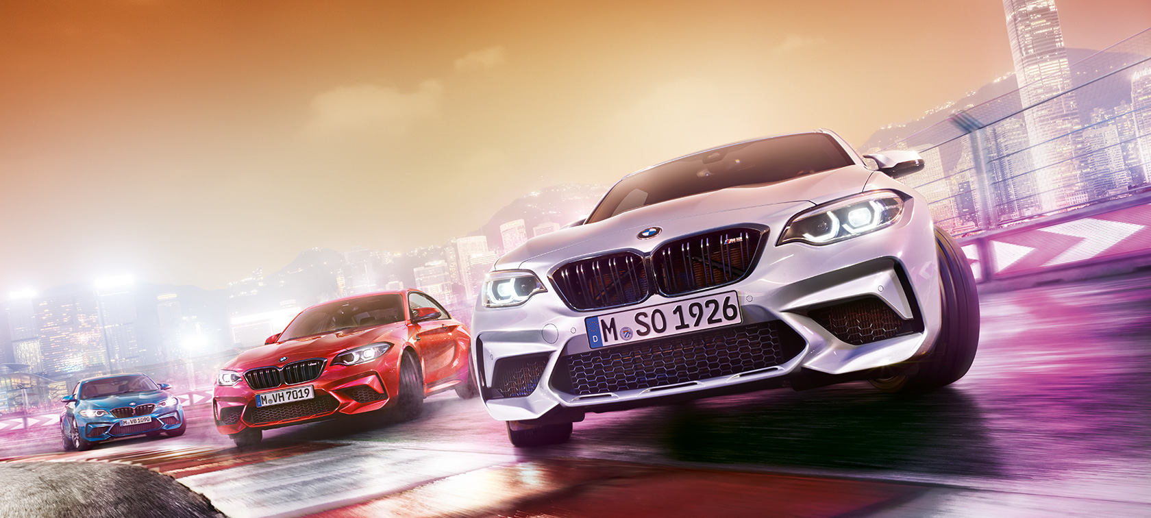 BMW M2 Competition w akcji. Trzy BMW M2 Competition driftują na torze wyścigowym z widokiem na nocny miejski pejzaż.