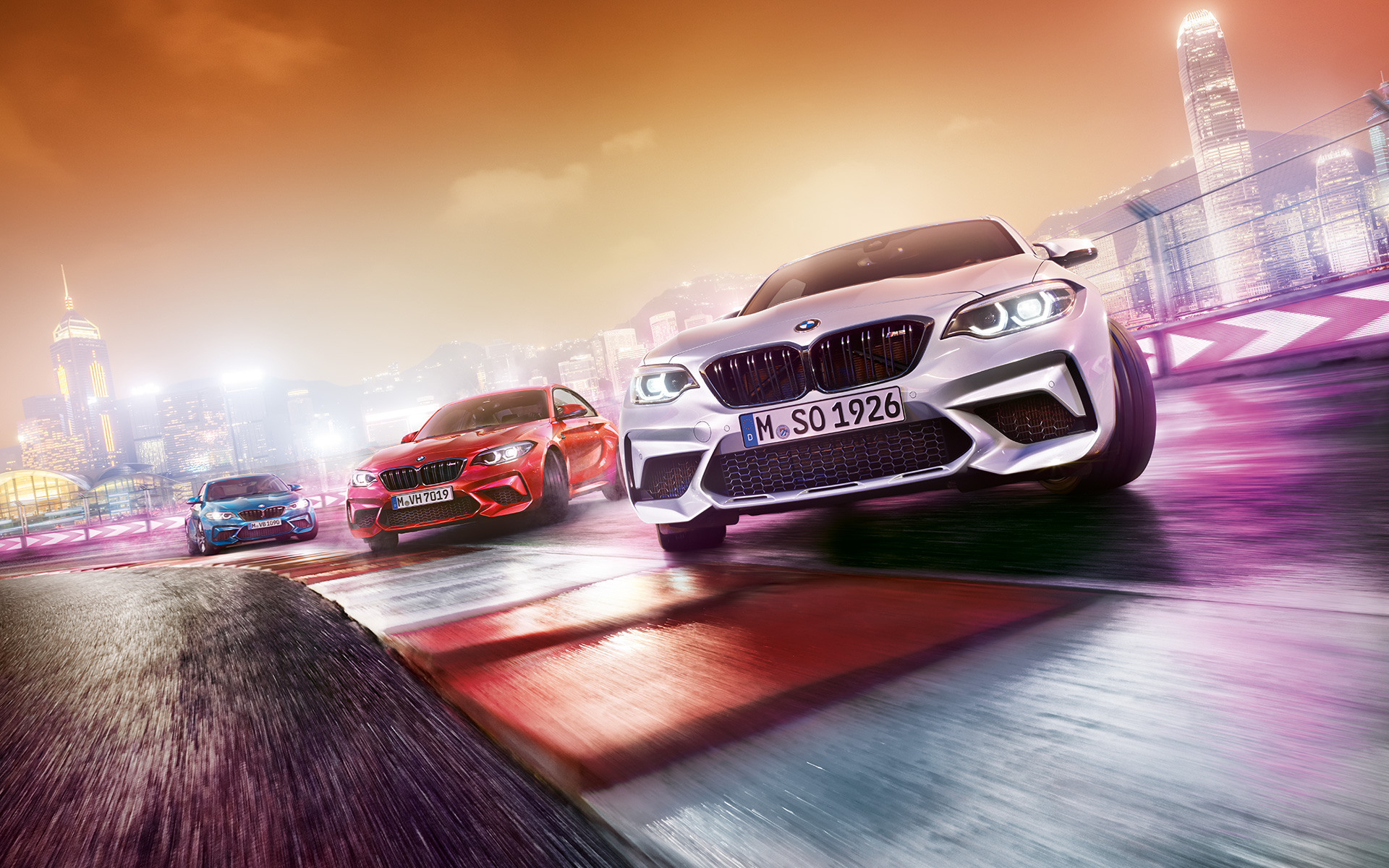 BMW M2 Competition w akcji. Trzy BMW M2 Competition driftują na torze wyścigowym w nocym miejskim pejzażu.