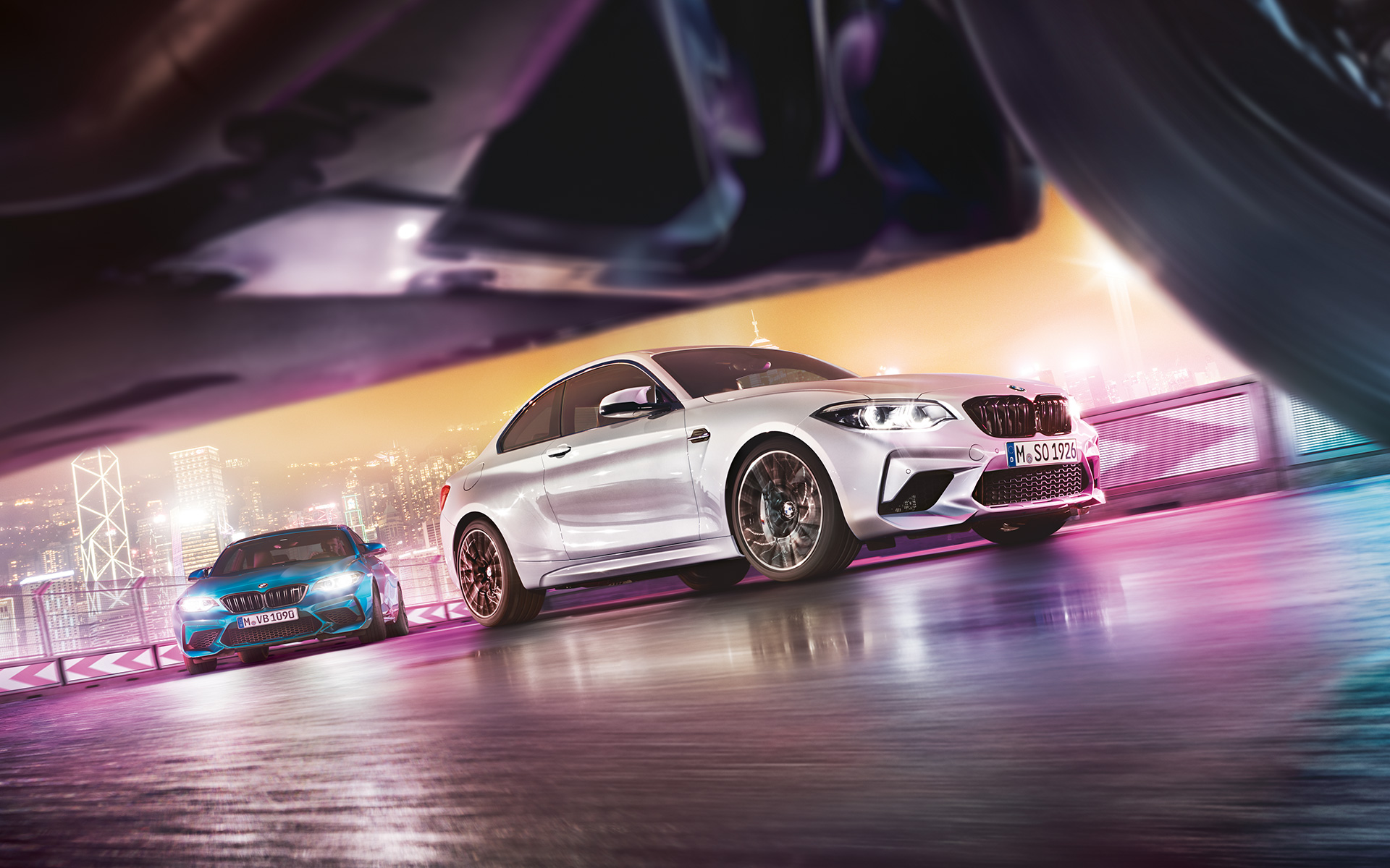 BMW M2 Competition na rozświetlonym asfalcie. Dwa BMW M2 Competition na torze wyścigowym przed nocnym miejskim pejzażem z widokiem z perspektywy ulicy.