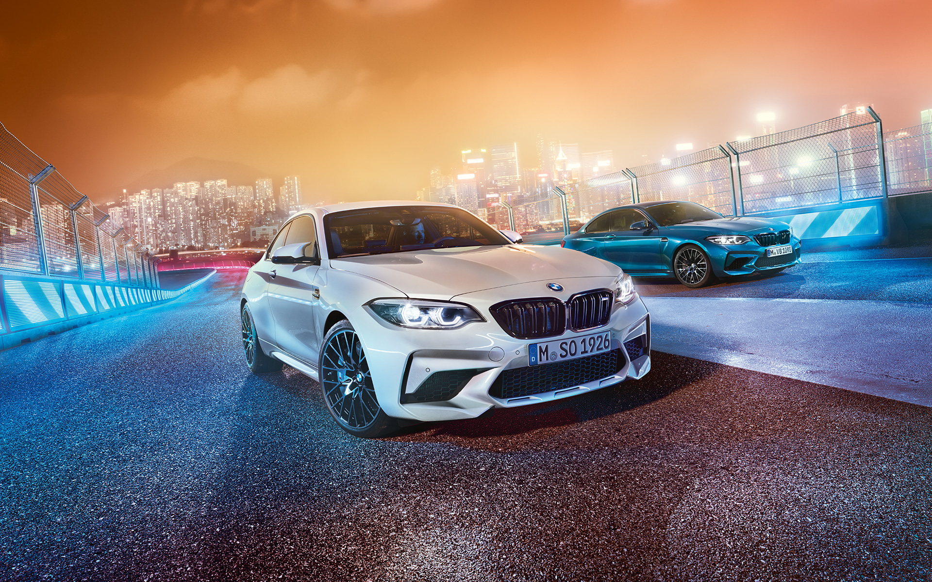 Dwa BMW M2 Competiotion na torzy wyścigowym. Białe i niebieskie BMW M2 Competition rzut z nachylonej perspektywy z nocnym miejskim pejzażem w tle.