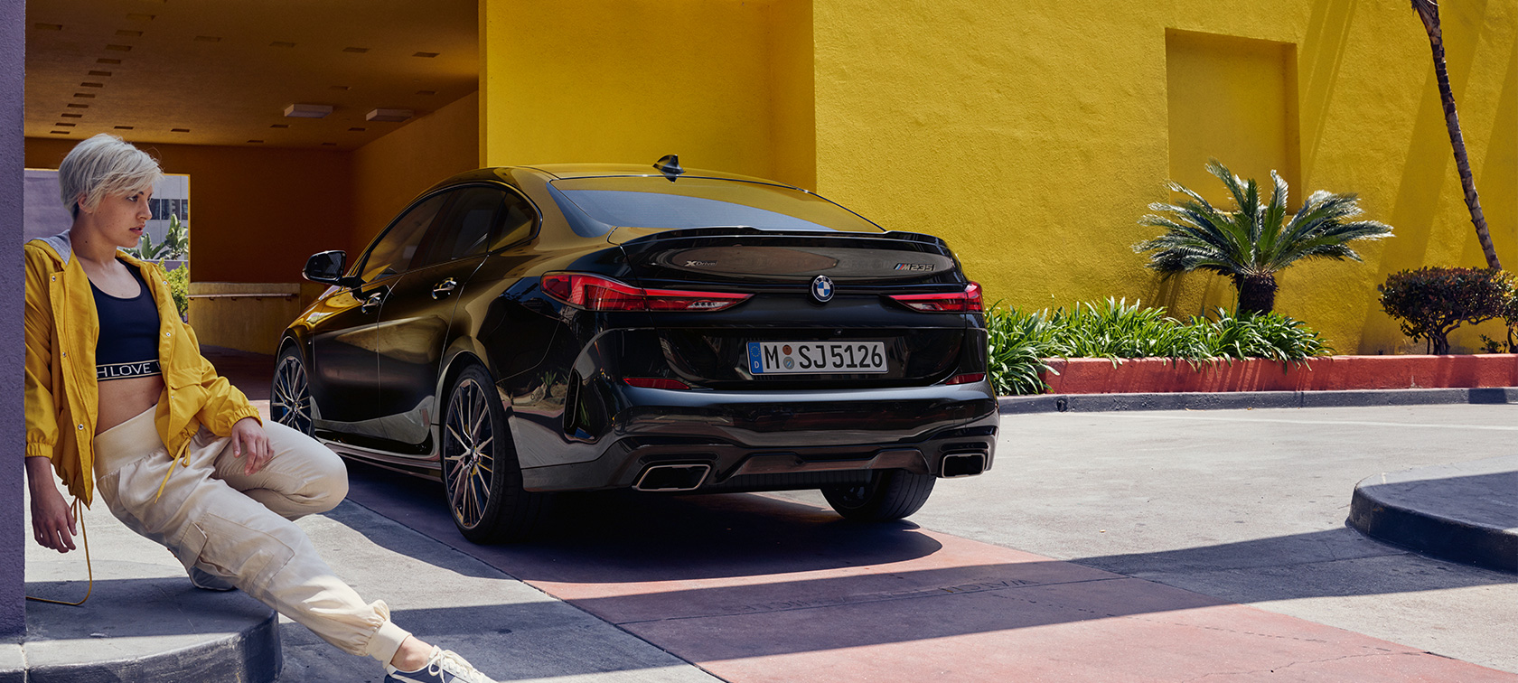 Tylny półprofil BMW M235i xDrive Gran Coupé BMW M235i xDrive Gran Coupé szafirowoczarny metalizowany F44 tylny półprofil z kobietą na pierwszym planie
