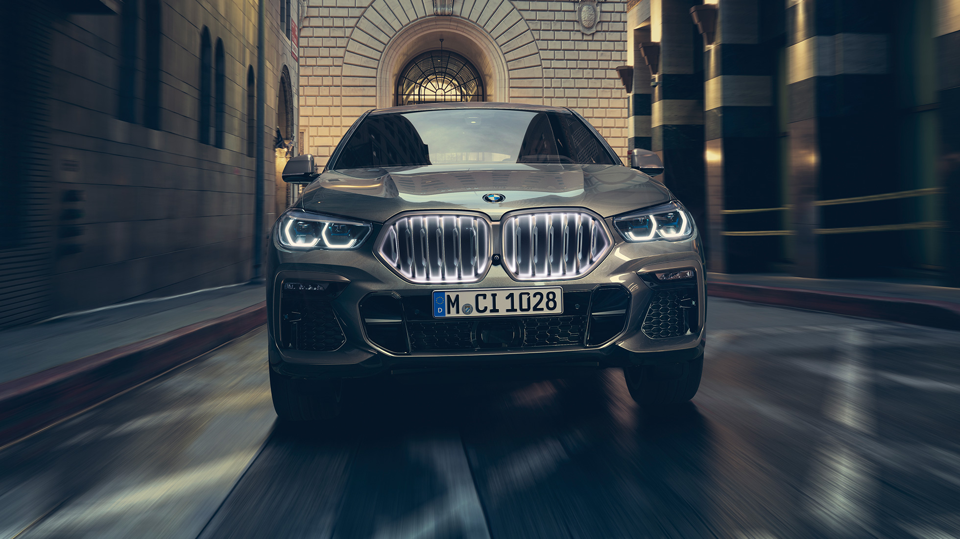 BMW X6 M50i z atrapą chłodnicy BMW z efektem „Iconic Glow”. BMW X6 M50i, ujęcie w perspektywie przed scenerią miejską.