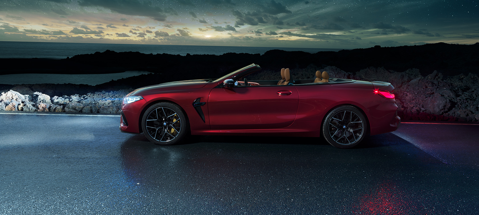 BMW M8 Competition Cabrio, widok z boku. BMW M8 Competition Cabrio w metalizowanym kolorze czerwieni Motegi z otwartym dachem, widok z boku, stojące pod rozgwieżdżonym niebem na drodze biegnącej wzdłuż wybrzeża.