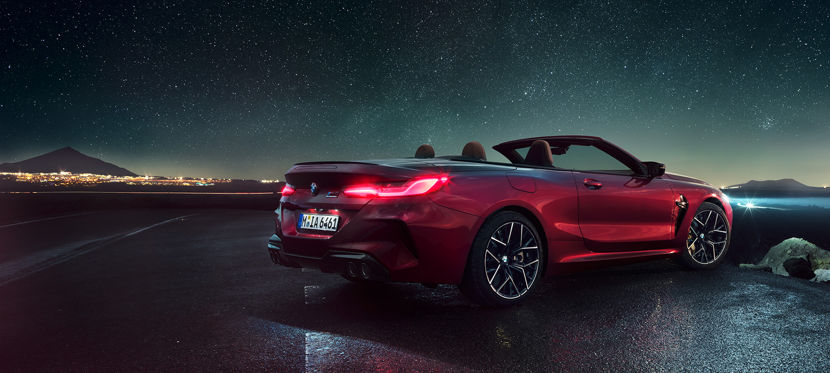 BMW M8 Competition Cabrio, tylny półprofil. BMW M8 Competition Cabrio w metalizowanym kolorze czerwieni Motegi z otwartym dachem, tylny półprofil, stojące pod rozgwieżdżonym niebem.