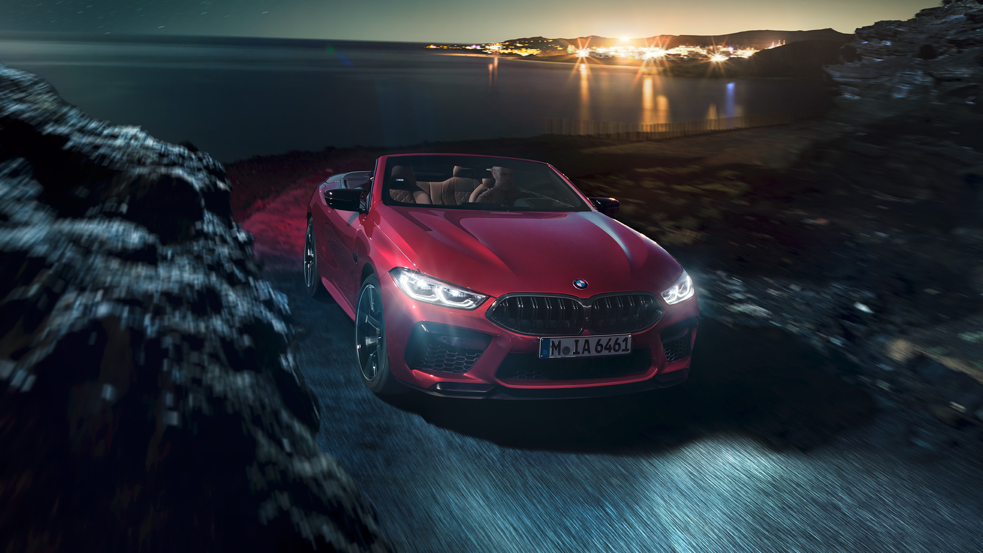 BMW M8 Competition Cabrio, przedni półprofil. BMW M8 Competition Cabrio w metalizowanym kolorze czerwieni Motegi z otwartym dachem, przedni półprofil, jadące w nocy drogą wzdłuż wybrzeża.