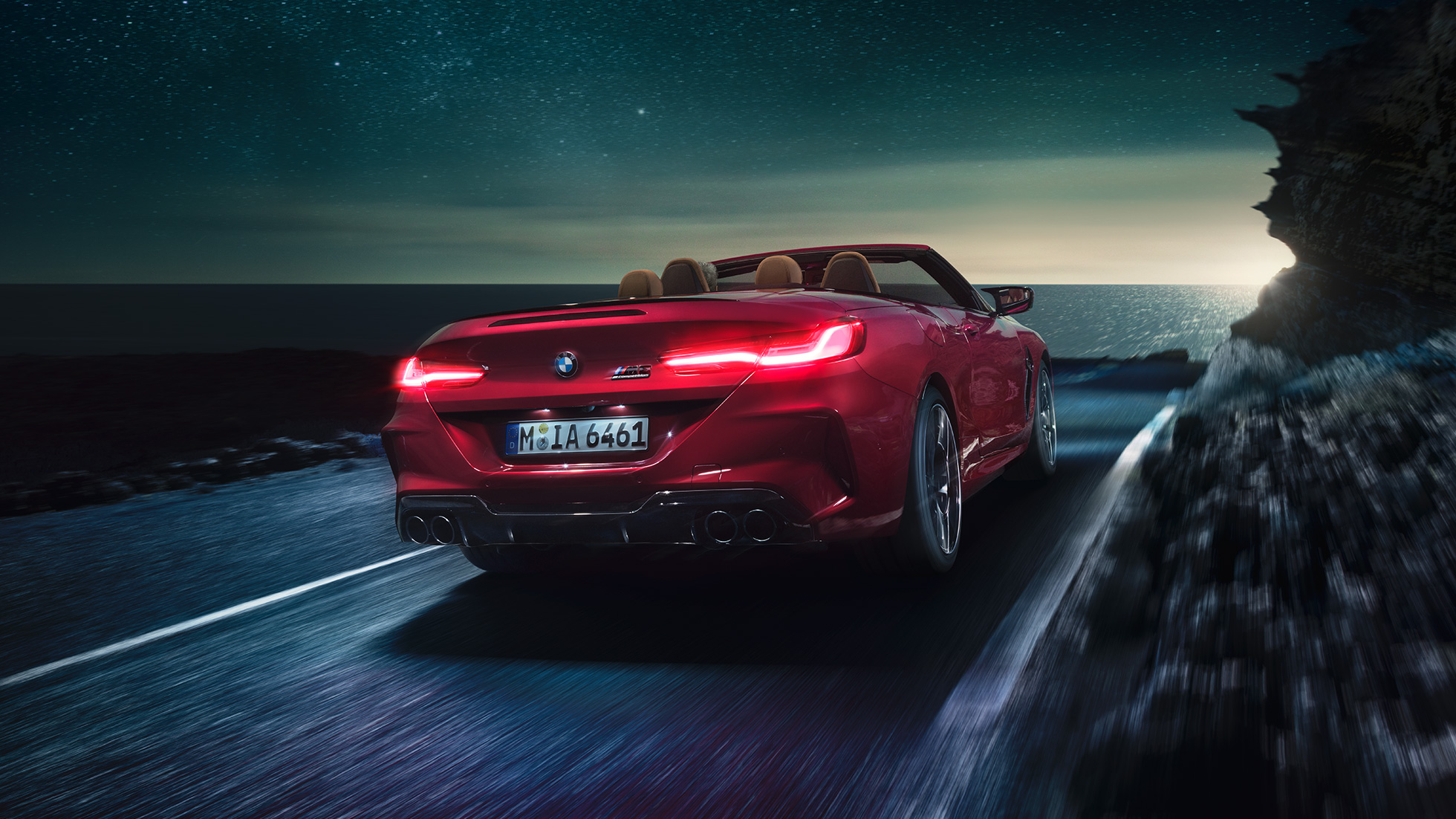 BMW M8 Competition Cabrio, tylny półprofil. BMW M8 Competition Cabrio w metalizowanym kolorze czerwieni Motegi z otwartym dachem, tylny półprofil, jadące w nocy drogą wzdłuż wybrzeża.