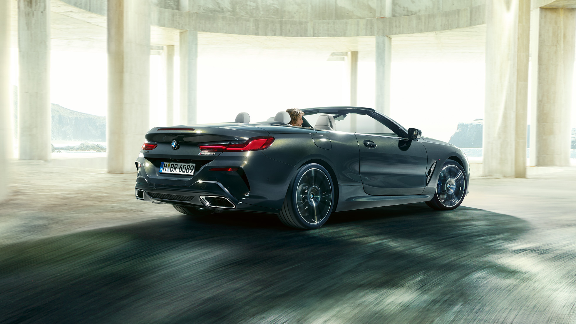 BMW M850i xDrive Cabrio, tylny półprofil. BMW M850i xDrive Cabrio w metalizowanym kolorze szarego drawitu z otwartym dachem, tylny półprofil, jadące przez betonową budowlę.