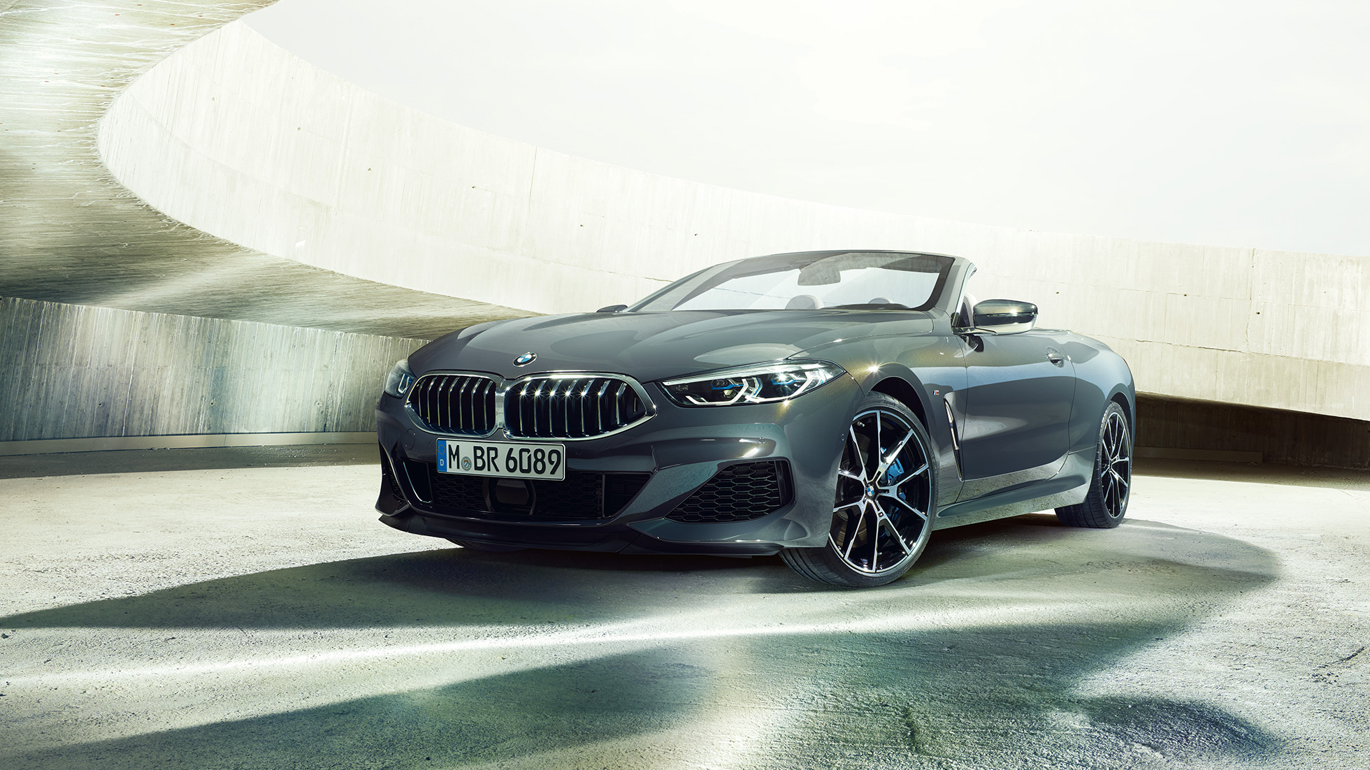 BMW M850i xDrive Cabrio, przedni półprofil. BMW M850i xDrive Cabrio w metalizowanym kolorze szarego drawitu z otwartym dachem, przedni półprofil, stojące w betonowej budowli.