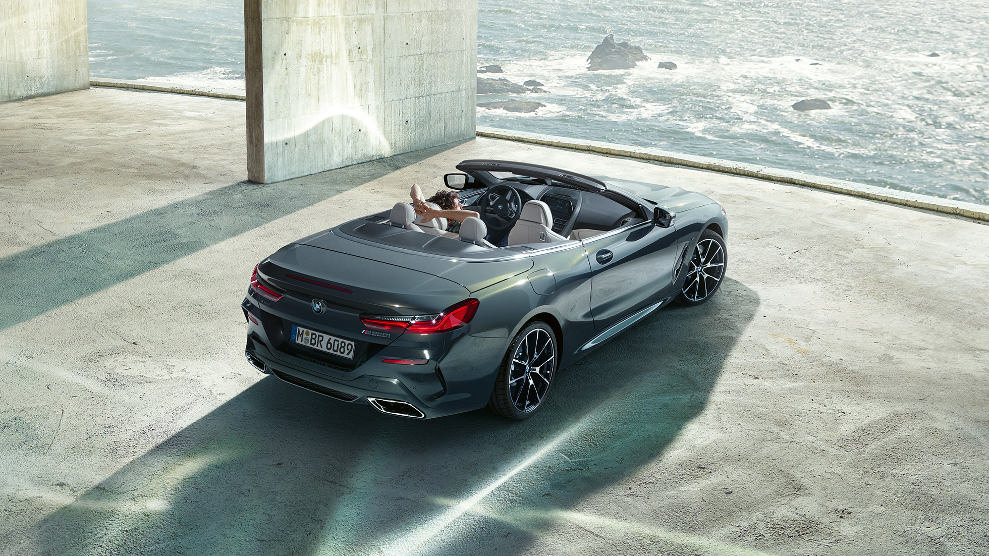 BMW M850i xDrive Cabrio, widok z góry. BMW M850i xDrive Cabrio w metalizowanym kolorze szarego drawitu z otwartym dachem, widok z góry, zaparkowane w metalowej budowli na tle oceanu.