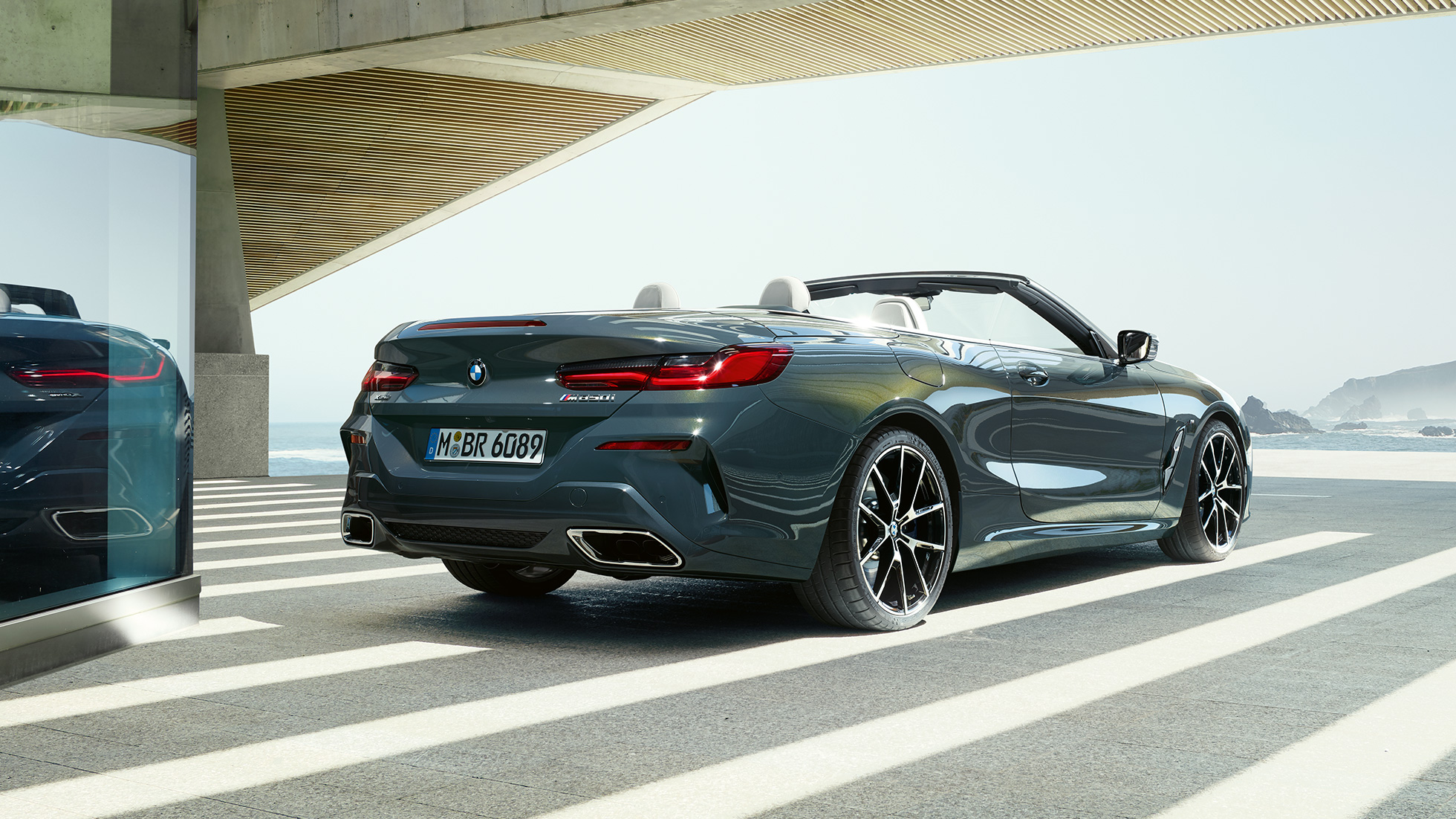 BMW M850i xDrive Cabrio, tylny półprofil. BMW M850i xDrive Cabrio w metalizowanym kolorze szarego drawitu z otwartym dachem, tylny półprofil, zaparkowane z widokiem na ocean.