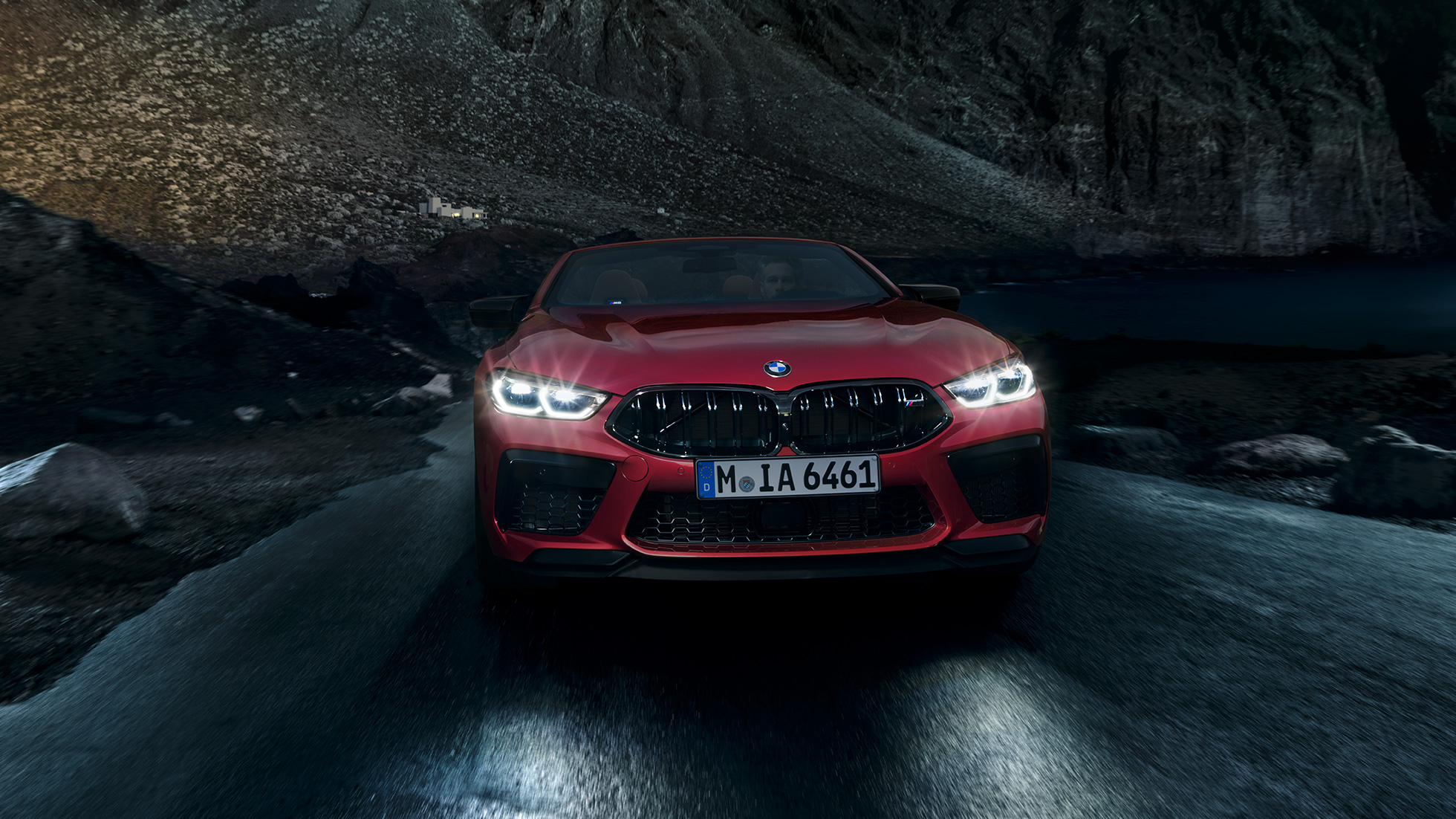BMW M8 Competition Cabrio, widok z przodu. BMW M8 Competition Cabrio w metalizowanym kolorze czerwieni Motegi ze świecącymi reflektorami, widok z przodu, jadące w nocy.