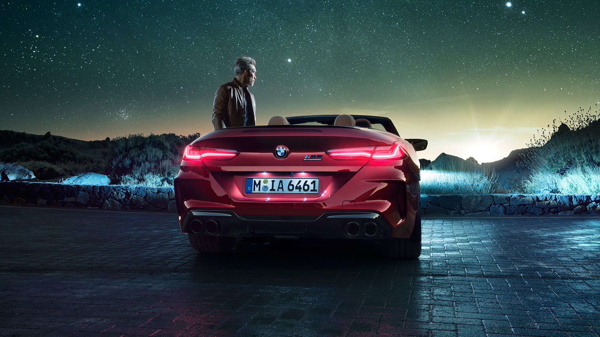 BMW M8 Competition Cabrio, widok z tyłu. BMW M8 Competition Cabrio w metalizowanym kolorze czerwieni Motegi z otwartym dachem, widok z tyłu, stojące wraz z mężczyzną pod rozgwieżdżonym niebem.