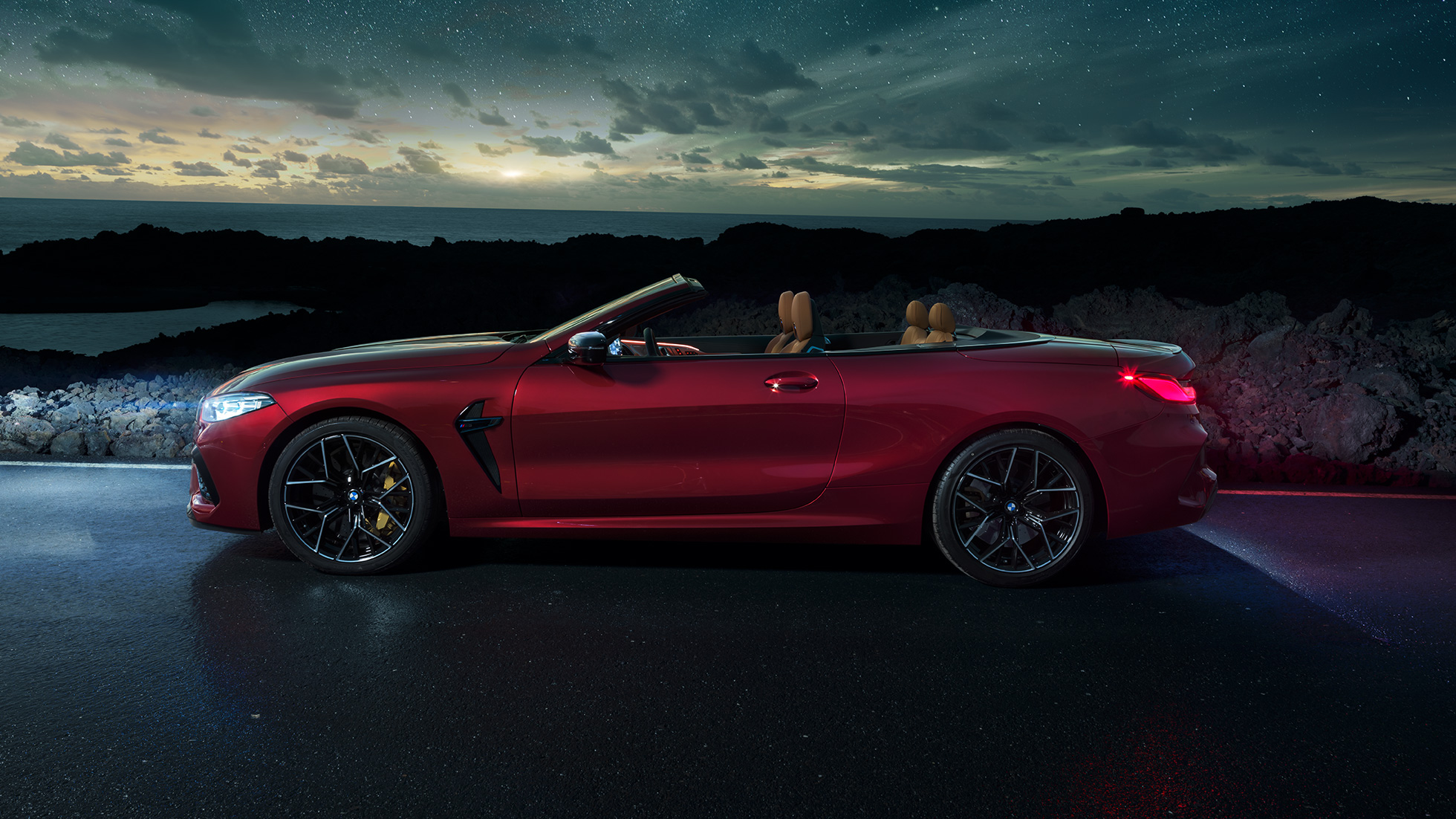 BMW M8 Competition Cabrio, widok z boku. BMW M8 Competition Cabrio w metalizowanym kolorze czerwieni Motegi z otwartym dachem, widok z boku, stojące w nocy pod rozgwieżdżonym niebem.