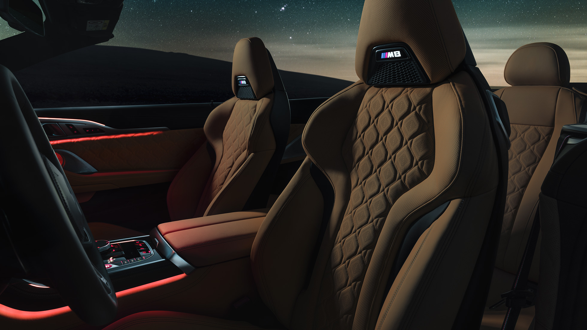 Fotele sportowe M obite skórą Merino. BMW M8 Competition Cabrio z otwartym dachem pod rozgwieżdżonym niebem, wnętrze z fotelami sportowymi M obitymi skórą Merino i czerwonym oświetleniem ambientowym.