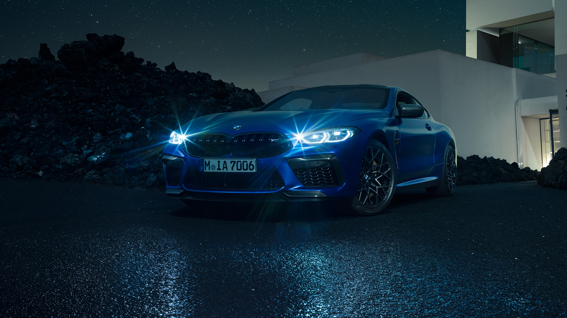 BMW M8 Competition Coupé, przedni półprofil. BMW M8 Competition Coupé w metalizowanym kolorze niebieskim Marina Bay ze świecącymi reflektorami LED, przedni półprofil w bezruchu, w nocy na tle willi.