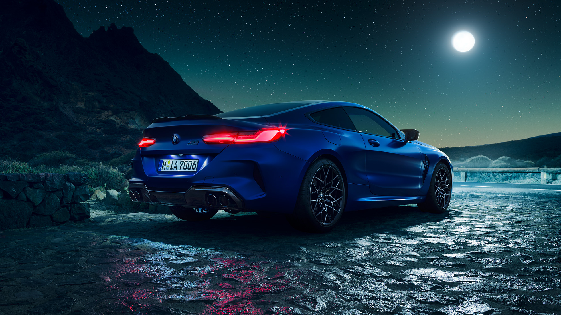BMW M8 Competition Coupé, tylny półprofil. BMW M8 Competition Coupé w metalizowanym kolorze niebieskim Marina Bay ze świecącymi reflektorami LED, tylny półprofil w bezruchu, przy księżycu w pełni.