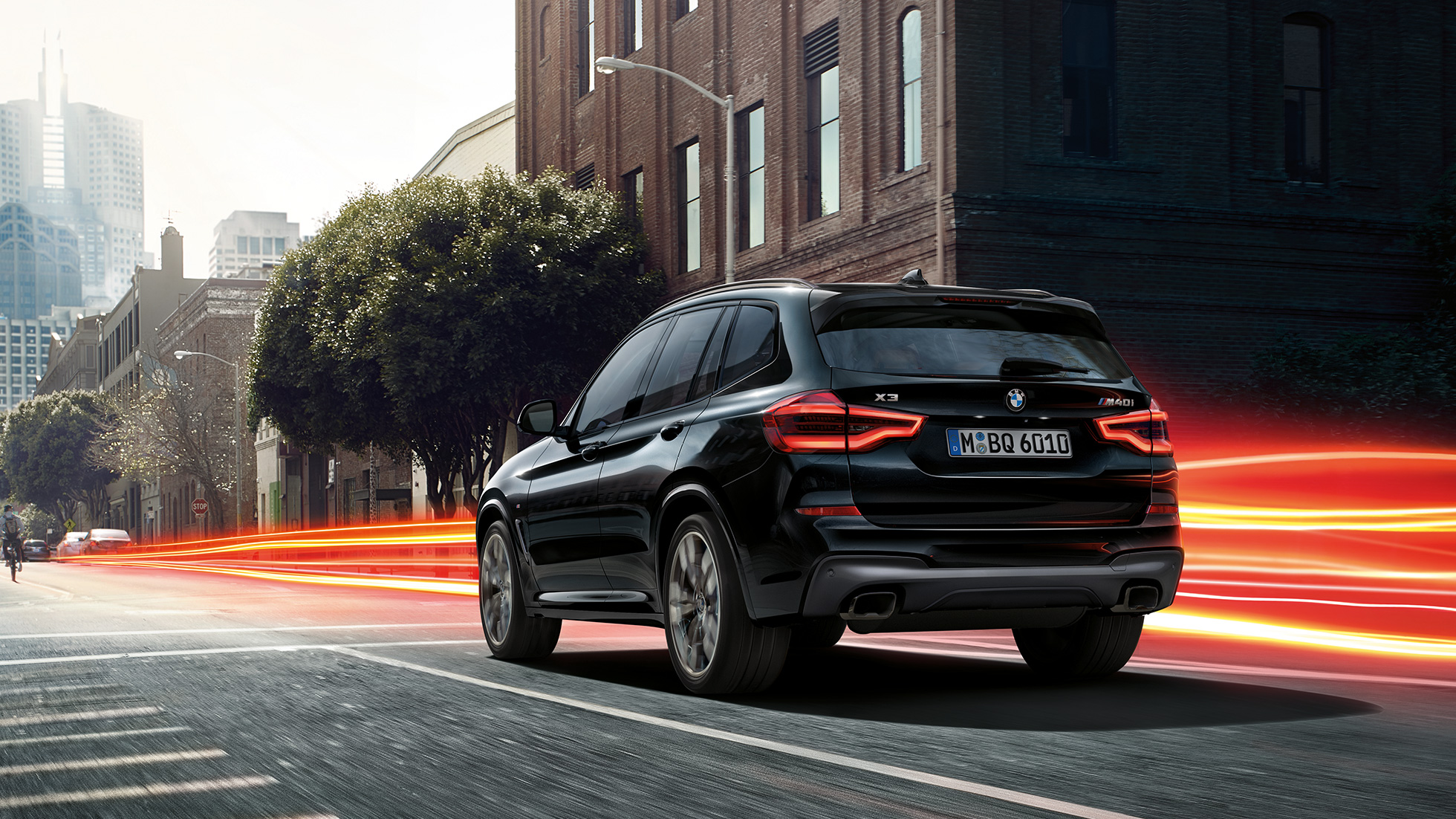 BMW X3 M40i, tylny półprofil. BMW X3 M40i, szafirowoczarny metalizowany, podczas jazdy, wygląd zewnętrzny, tylny półprofil na tle miasta z pasami świetlnymi.