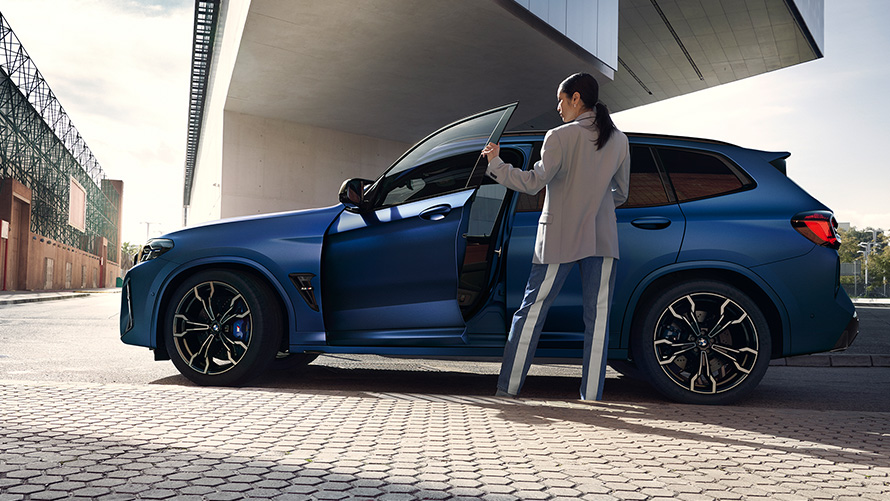 BMW X3 M Competition F97 LCI 2021 widok z boku z otwartymi drzwiami i modelką BMW X3 M Competition F97 LCI modernizacja 2021 mroźny niebieski Marina Bay metalizowany widok z boku z otwartymi drzwiami i modelką