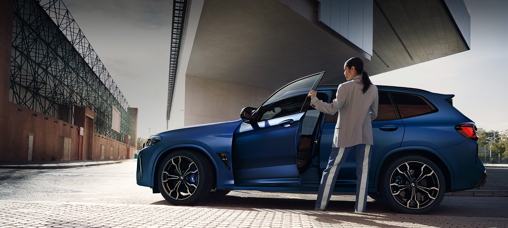 BMW X3 M Competition F97 LCI modernizacja 2021 widok z boku podczas postoju BMW X3 M Competition F97 LCI modernizacja 2021 mroźny niebieski Portimao BMW Individual metalizowany widok z boku podczas postoju z otwartymi drzwiami i modelką