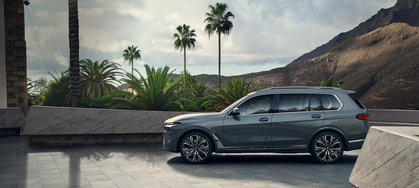 BMW X7 G07 LCI widok z boku połyskujący miedziany szary metalizowany BMW X7 G07 LCI widok z boku podczas postoju na tle drzew połyskujący miedziany szary metalizowany