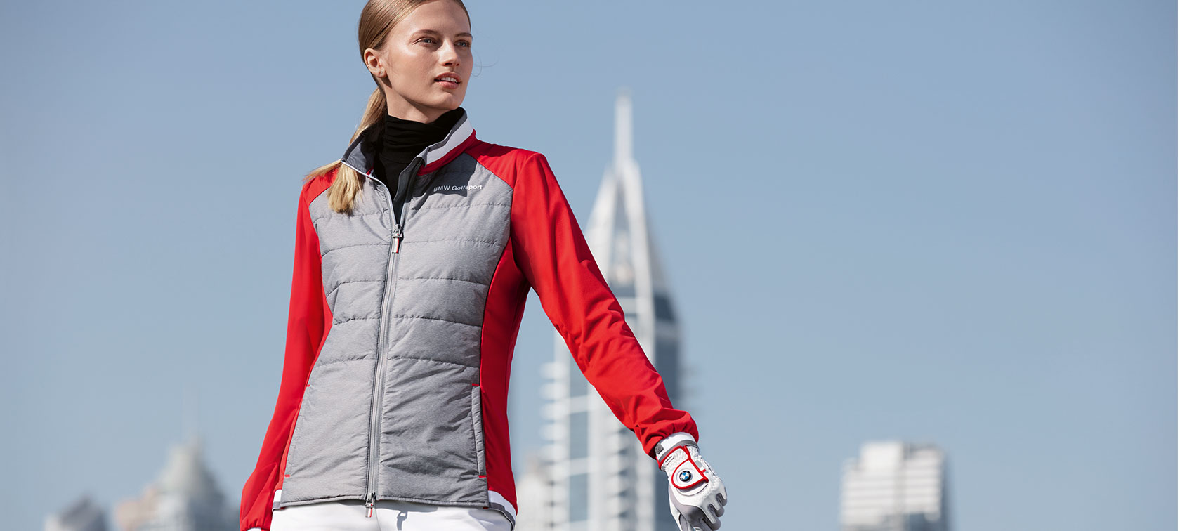 Damska kurtka BMW Golfsport. Na zdjęciu widać młodą kobietę prezentującą produkty premium z kolekcji BMW Golfsport na tle panoramy miasta.