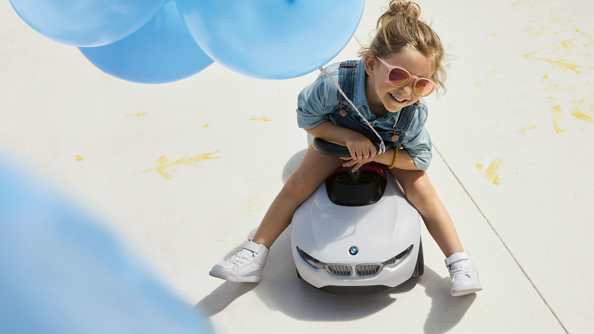 Kolekcja BMW Kids. Uśmiechnięta dziewczynka w różowych okularach i z niebieskim balonem w ręku siedzi na samochodziku BMW Baby Racer.