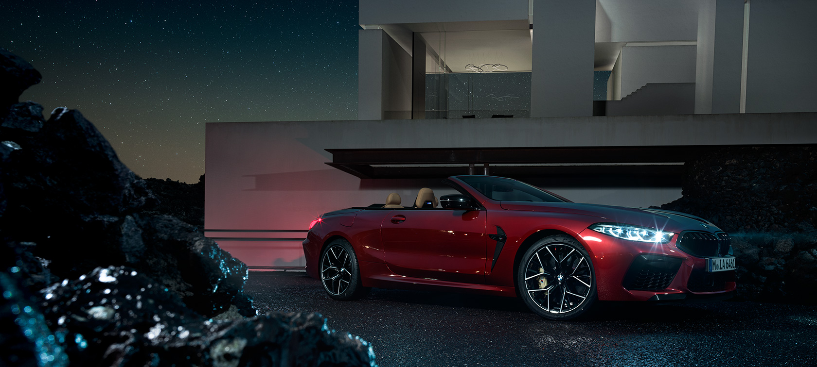 BMW M8 Competition Cabrio, boczny półprofil. BMW M8 Competition Cabrio w kolorze Motegi Red metallic. Ujęcie z bocznego półprofilu, stojące nocą na tle willi.