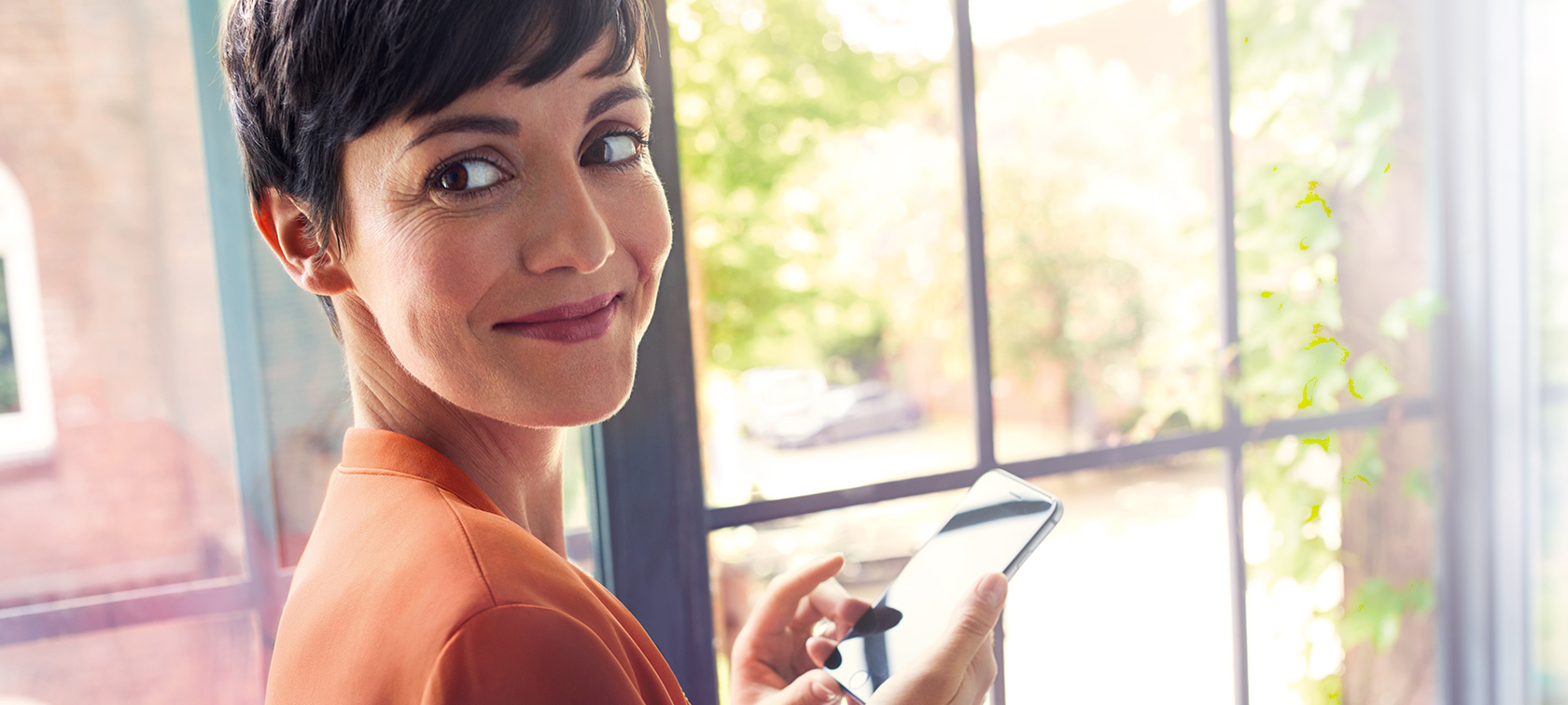 Brunetka ze smartfonem Brunetka w pomarańczowej kurtce stoi przy oknie ze smartfonem i uśmiecha się